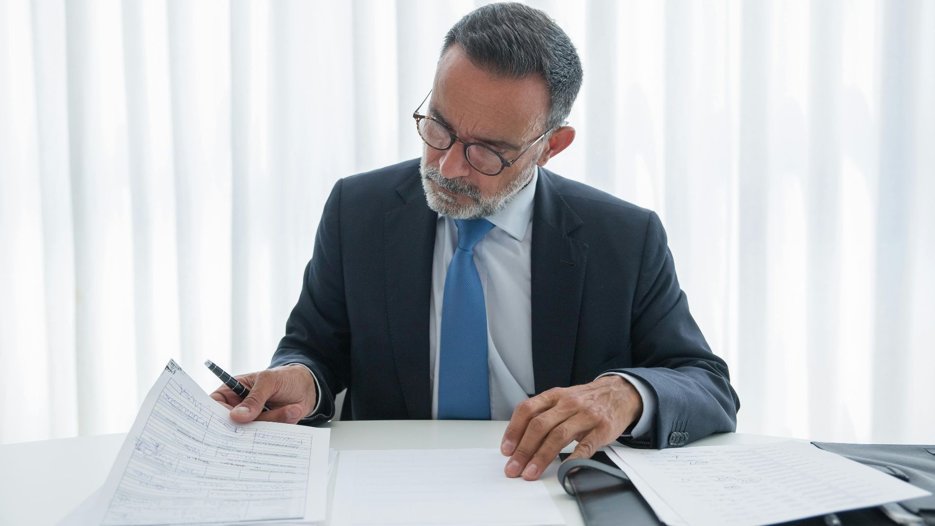 Elderly businessman in suit reviewing documents at desk. Professional setting.