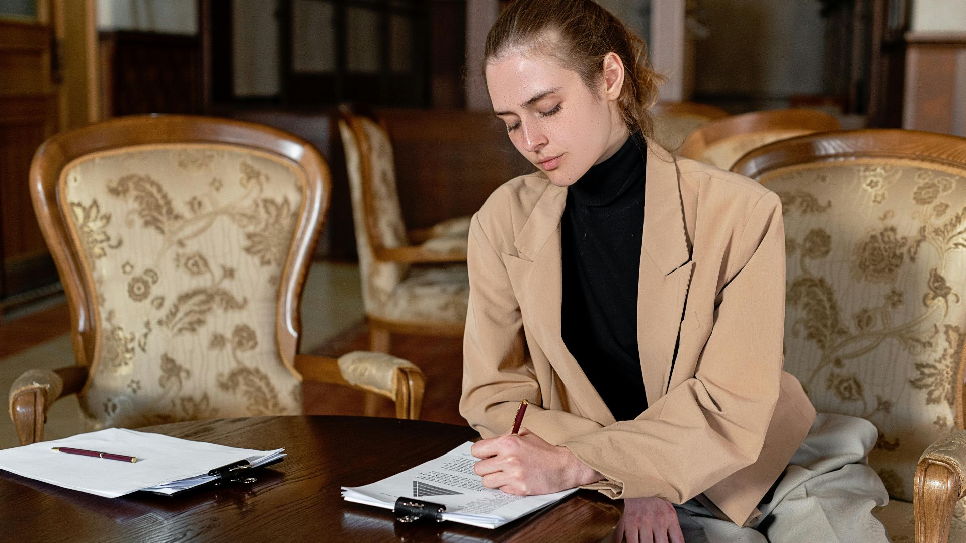 Young woman in a brown blazer signing documents at a table in a stylish office.