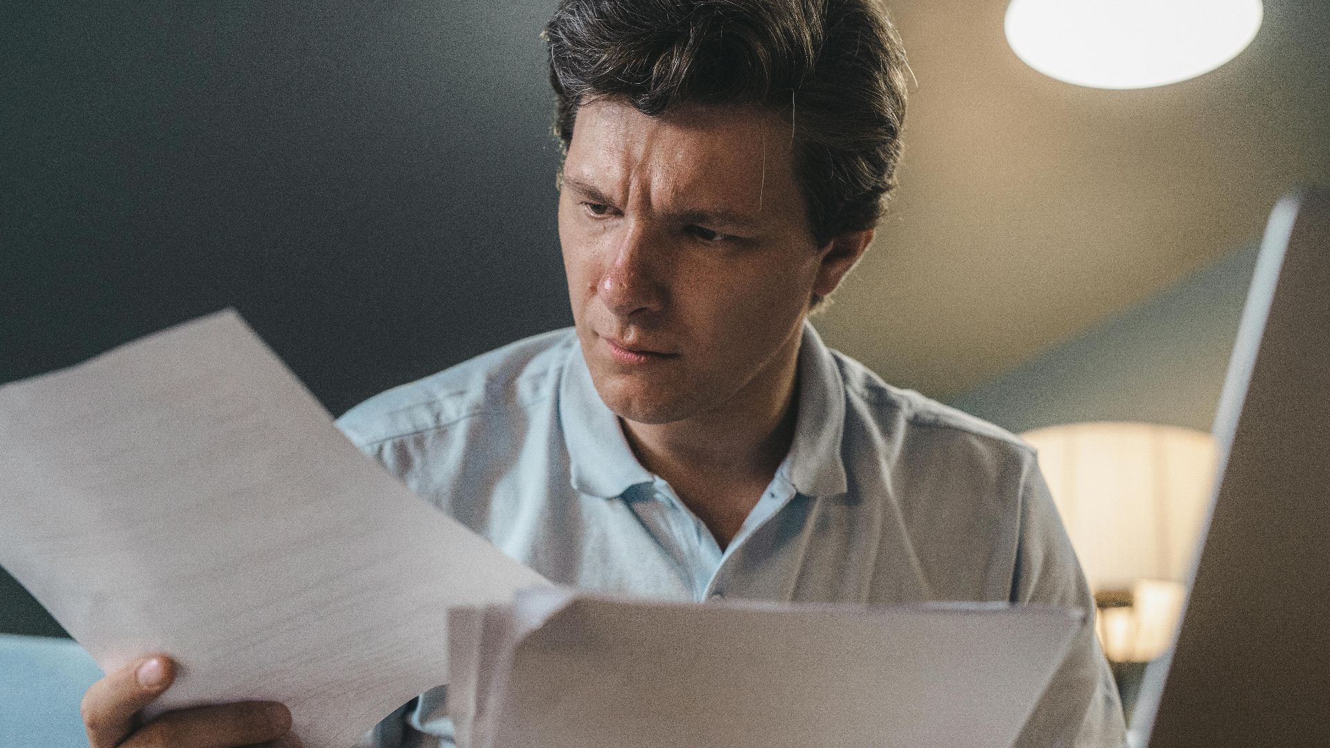 A man intently examines papers, seated indoors under warm lighting, focusing on his work.