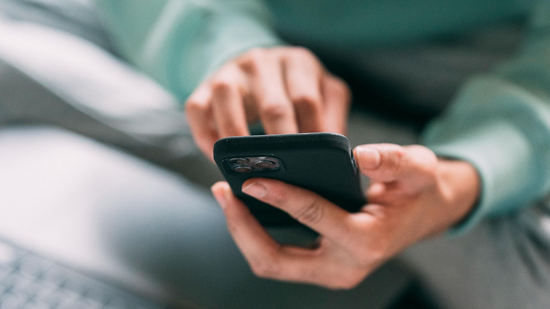 Crop anonymous male freelancer text messaging on modern cellphone while sitting on bed with laptop during remote work at home