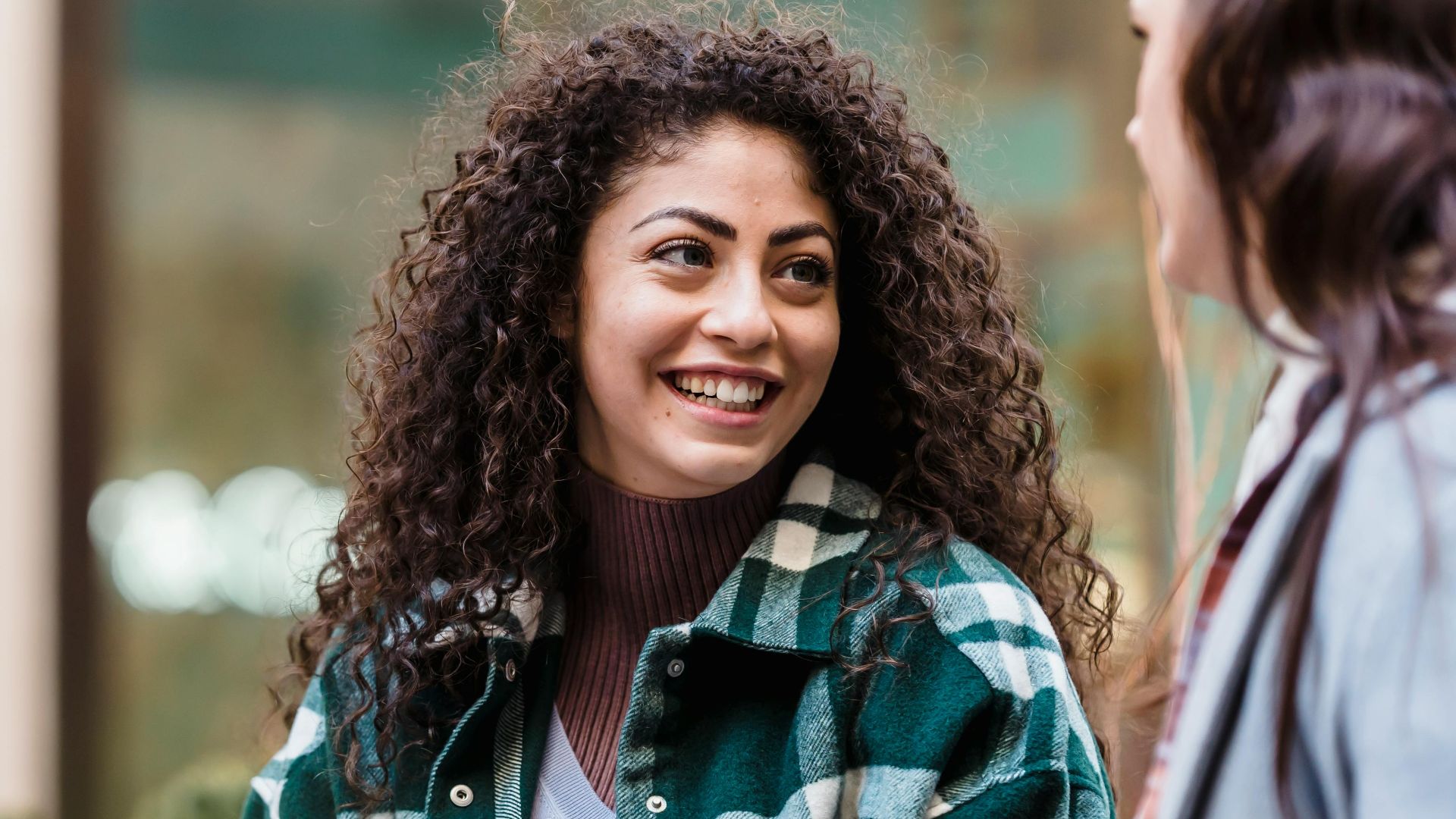 Cheerful woman with long curly hair in casual clothes standing and communicating with friend on street in daylight