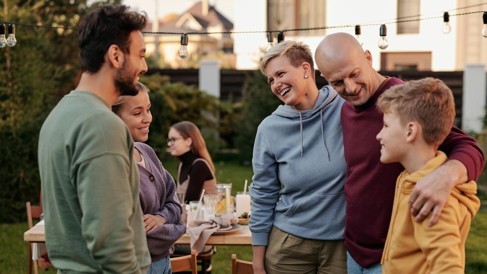 Happy family in casual clothes laughing and having picnic in backyard with string of light bulbs in daytime