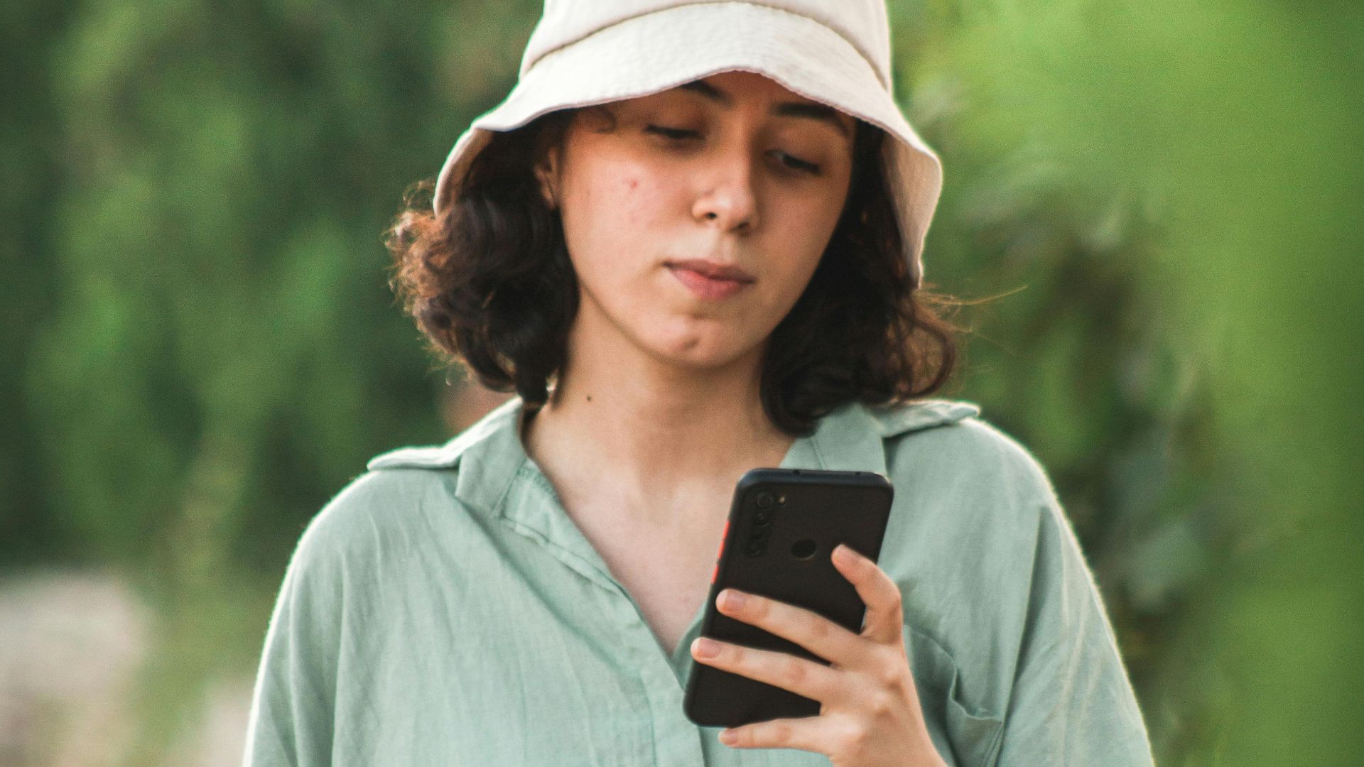 A young woman in a bucket hat uses her smartphone while walking outdoors.