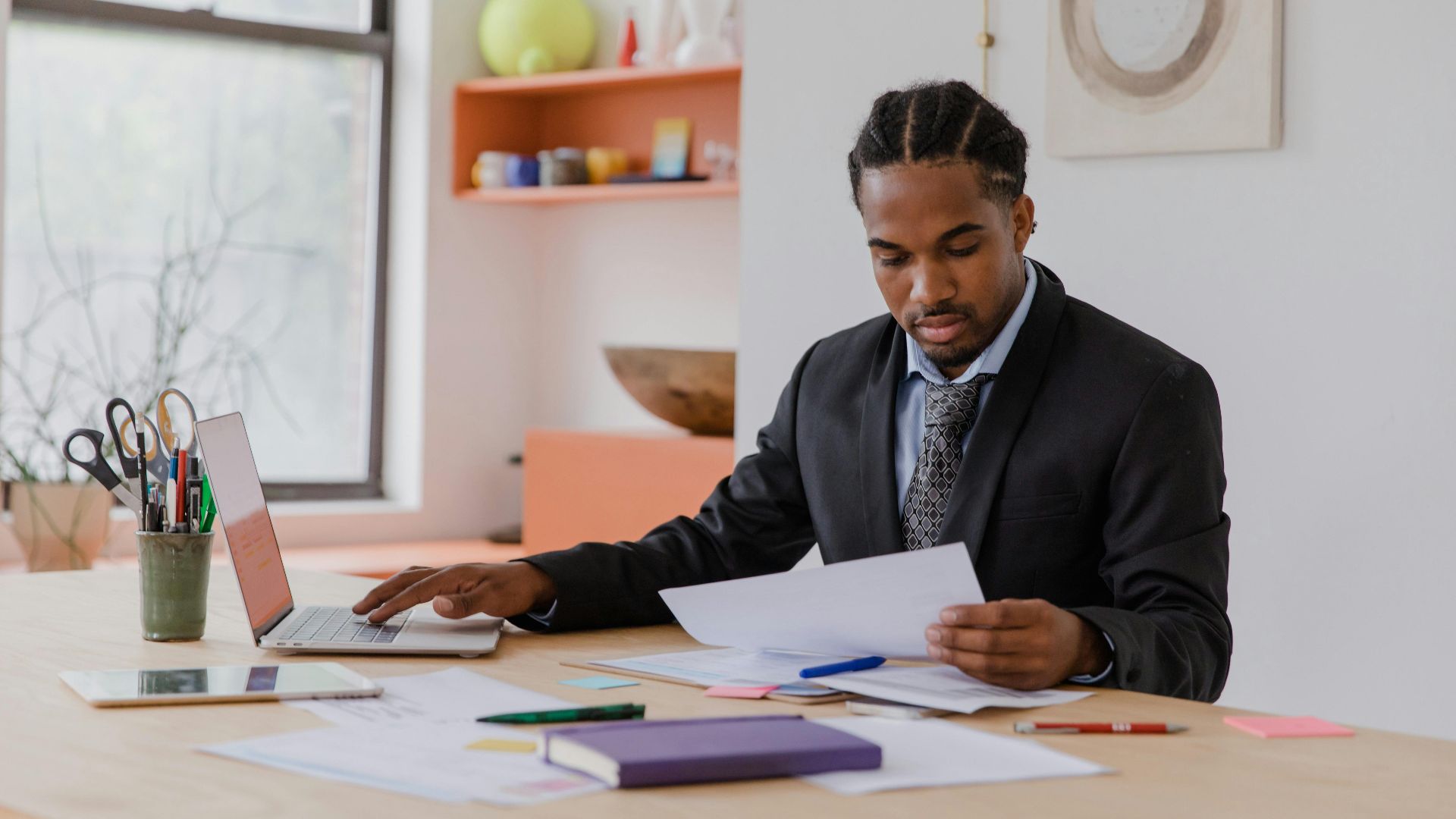 A professional businessman in a suit working at a desk with documents and a laptop in an office setting.