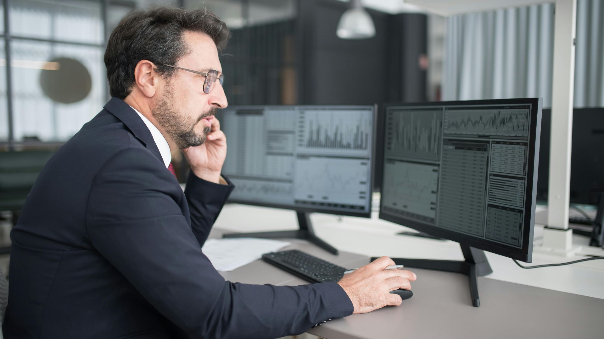 Businessman analyzing stock market data on dual monitors in a modern office setting.