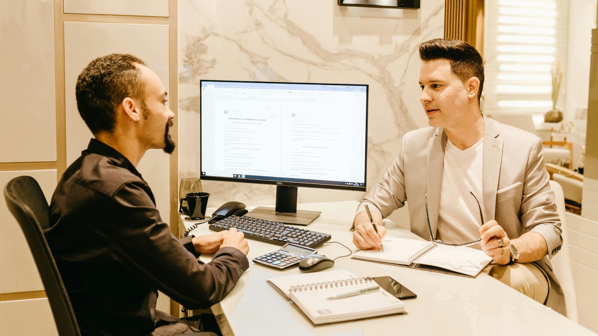 Two businessmen discussing work at a sleek and modern office desk, using computer and notes for collaboration.