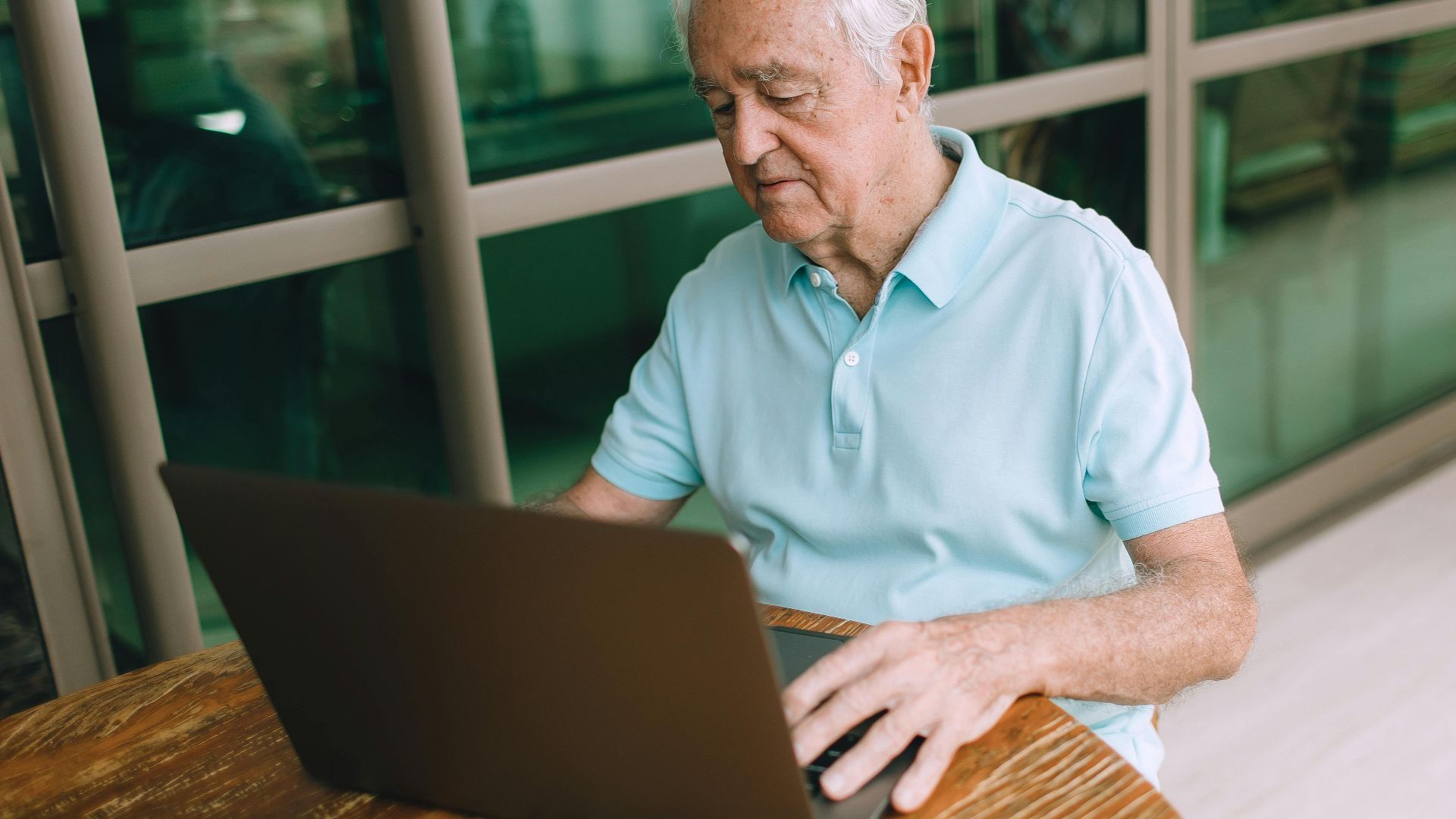 Elderly man sitting at a wooden table, working on a laptop indoors.
