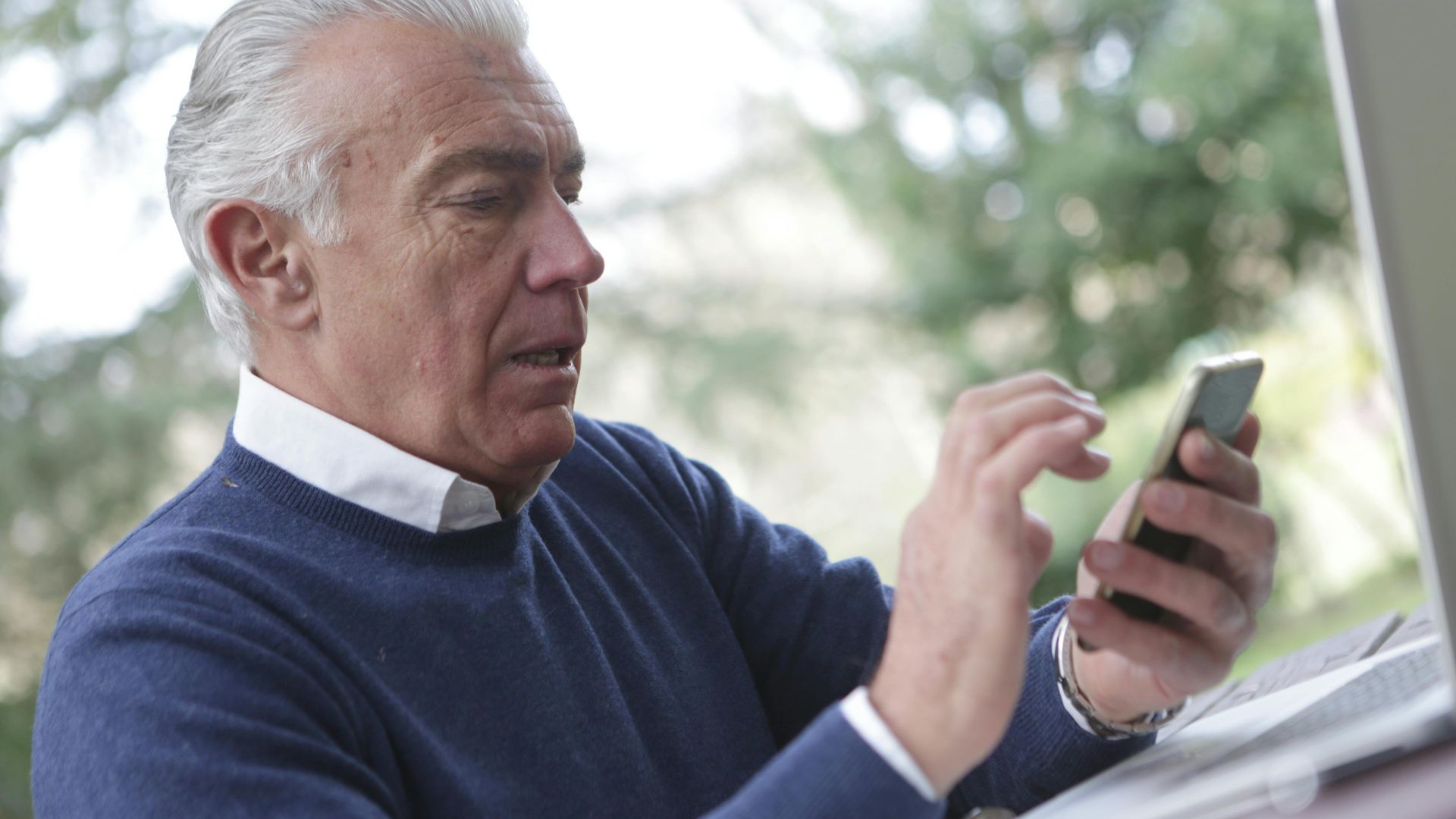 Elderly man using smartphone outdoors, working remotely on laptop.