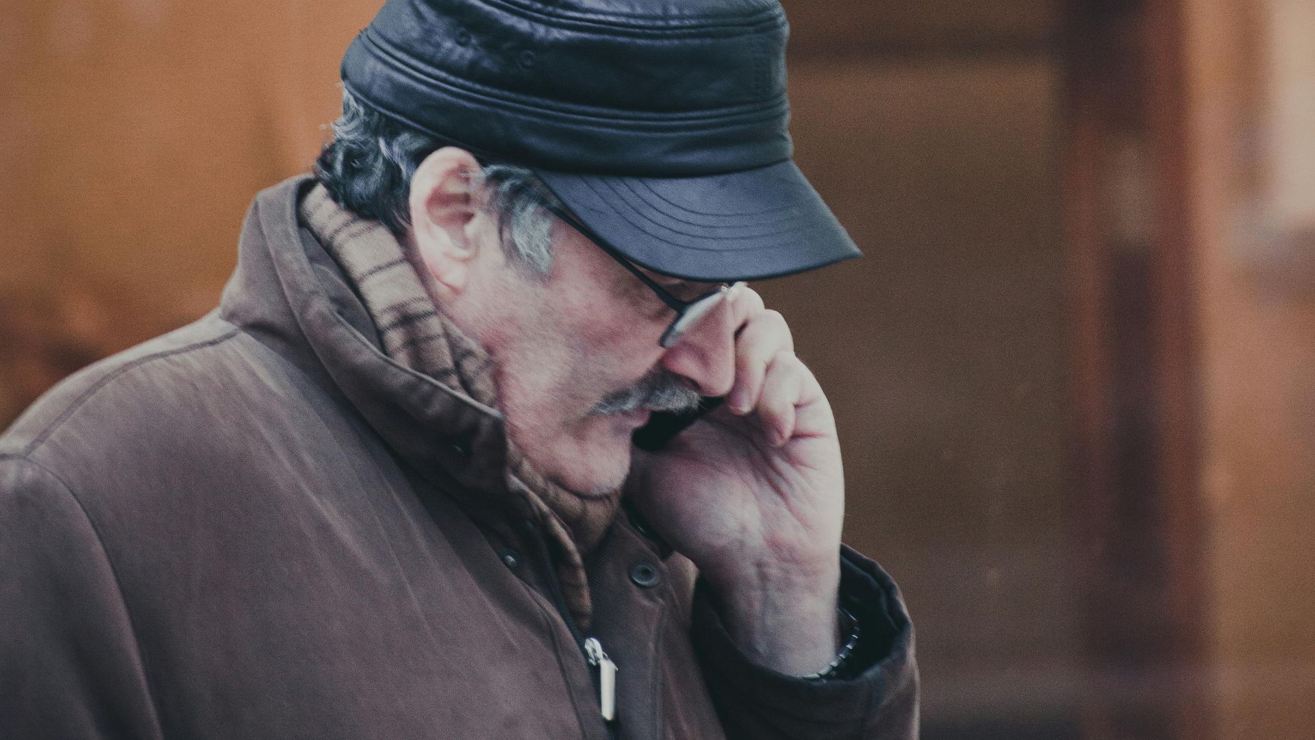 Senior man wearing a leather hat and coat, talking on the phone outdoors.
