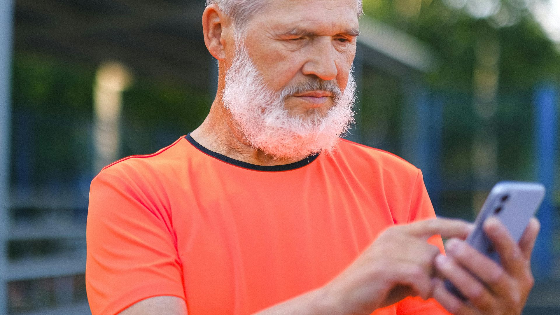 Crop elderly bearded male with gray hair browsing modern mobile phone while spending time in park