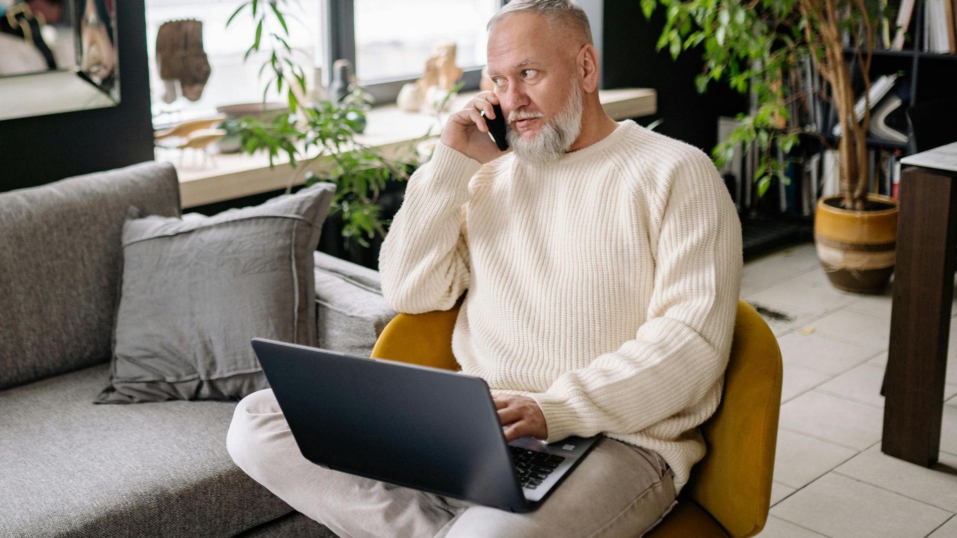 Elderly man multitasking at home, using laptop and phone, sitting comfortably indoors.