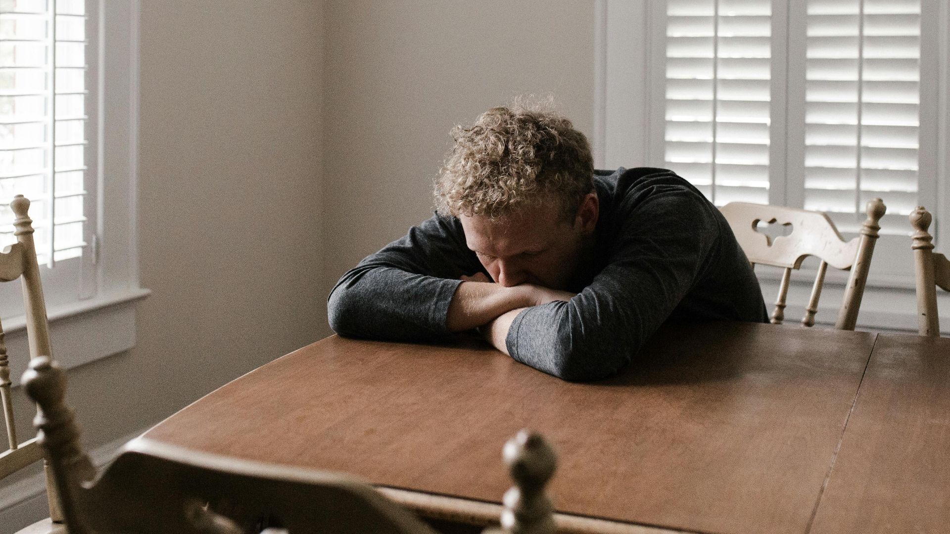 An adult man with emotions of sadness and anxiety sitting alone at a wooden table near windows.