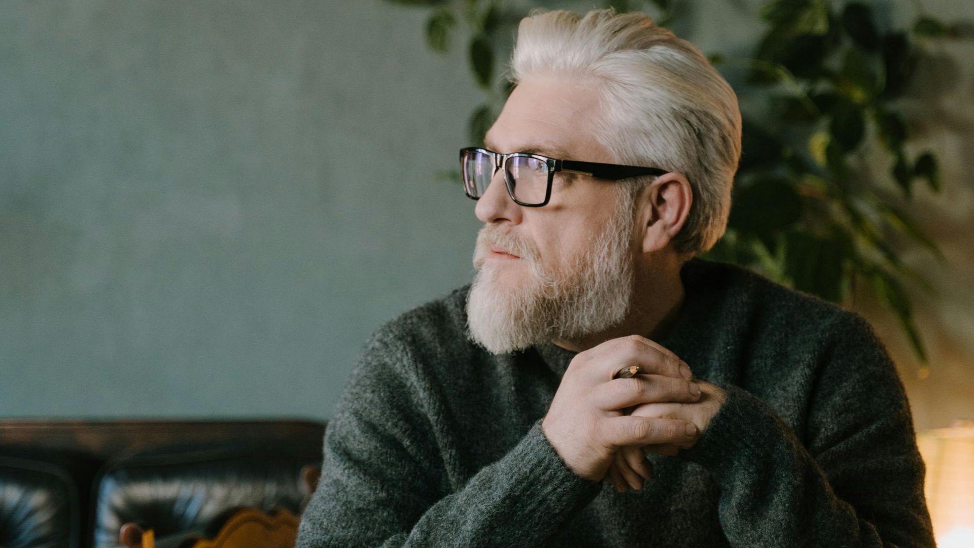Elderly man with white hair and glasses sits thoughtfully at a wooden desk in a cozy indoor setting.