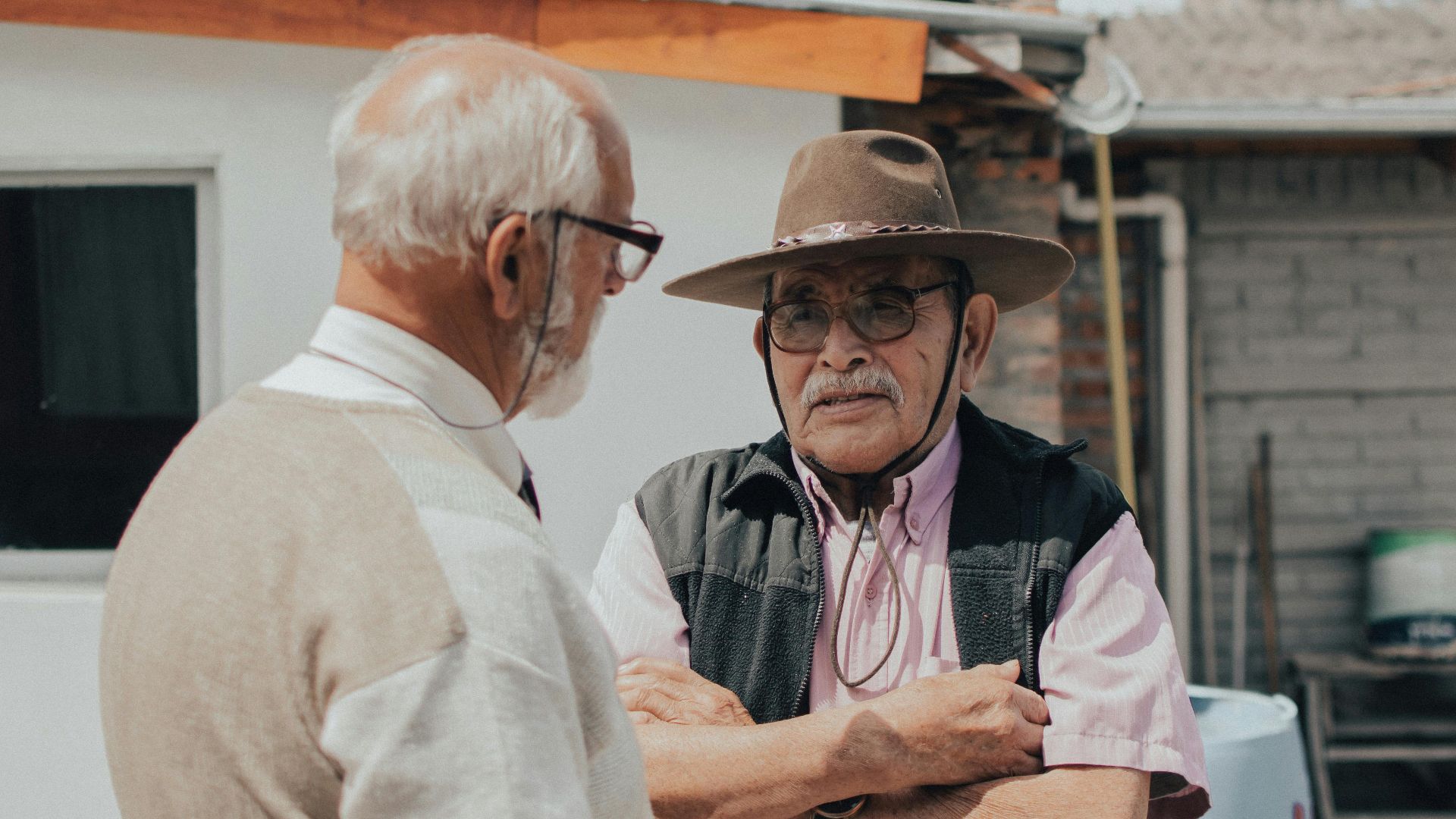 Two senior men engaged in conversation outside a rustic building on a sunny day.
