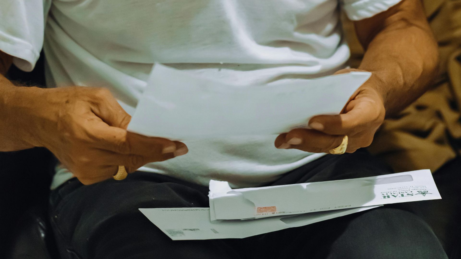 An elderly man in glasses reading papers, wearing a white t-shirt indoors.