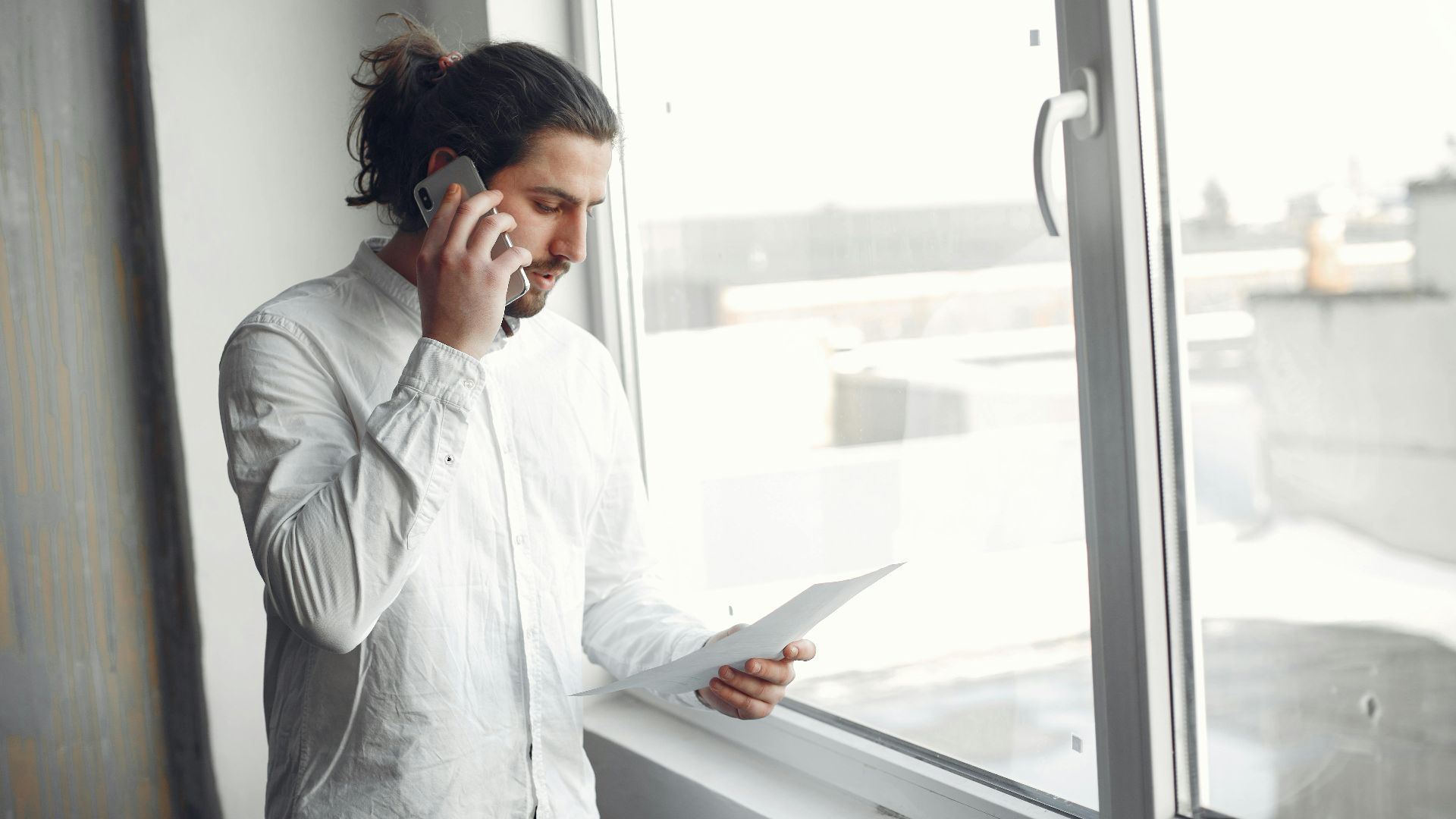 Young man in white shirt, on phone call holding a document, standing by a large window.