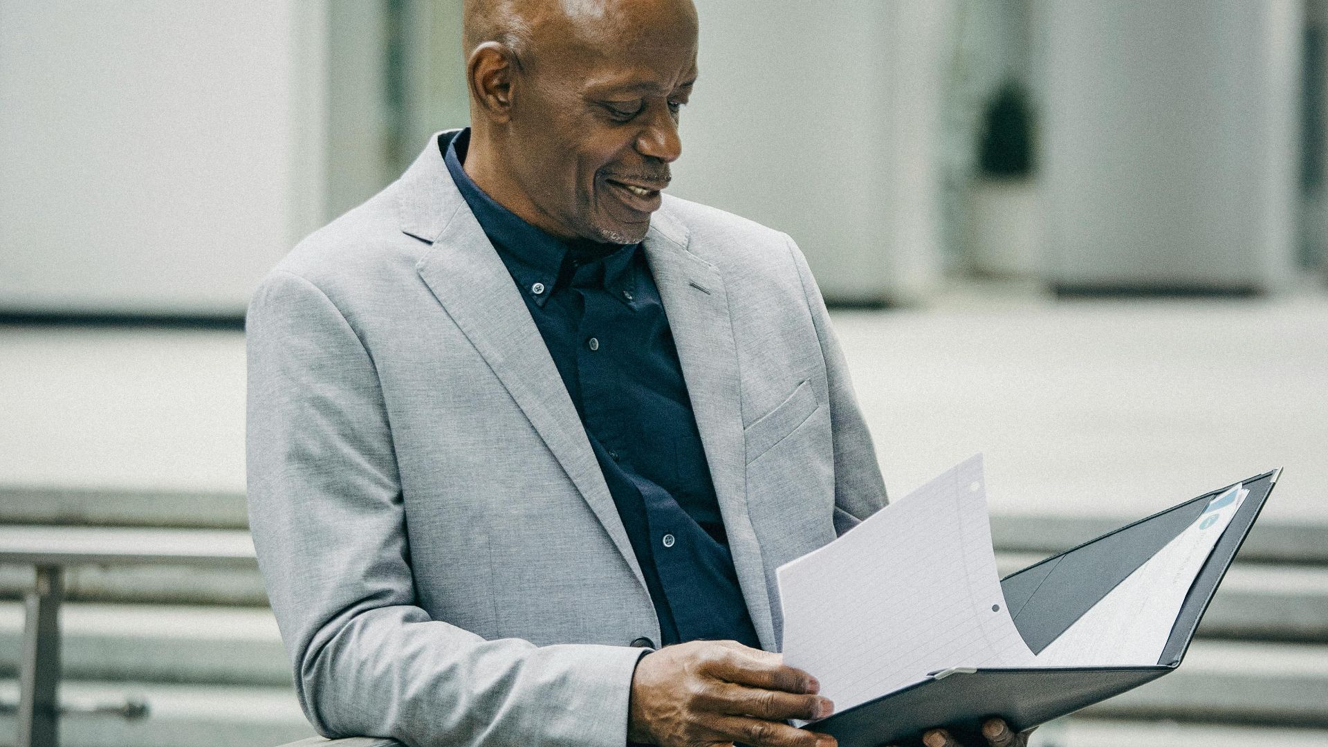 Content middle aged African American male manager in formal suit smiling while checking documents standing near modern building