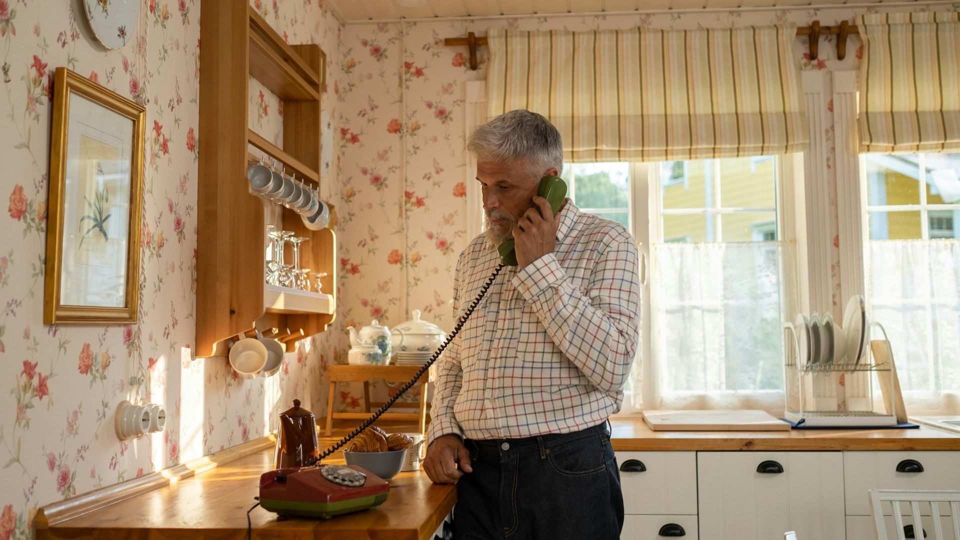 Senior man talks on a vintage rotary phone in a retro-styled kitchen with floral wallpaper and classic decor.