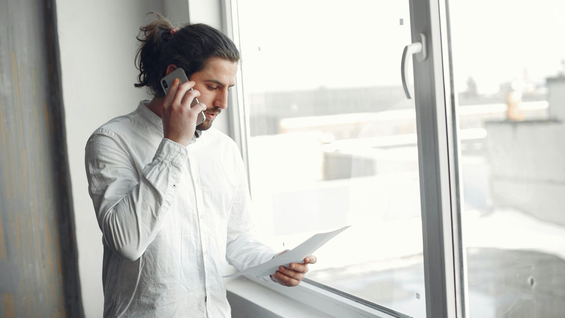 Young man in white shirt, on phone call holding a document, standing by a large window.