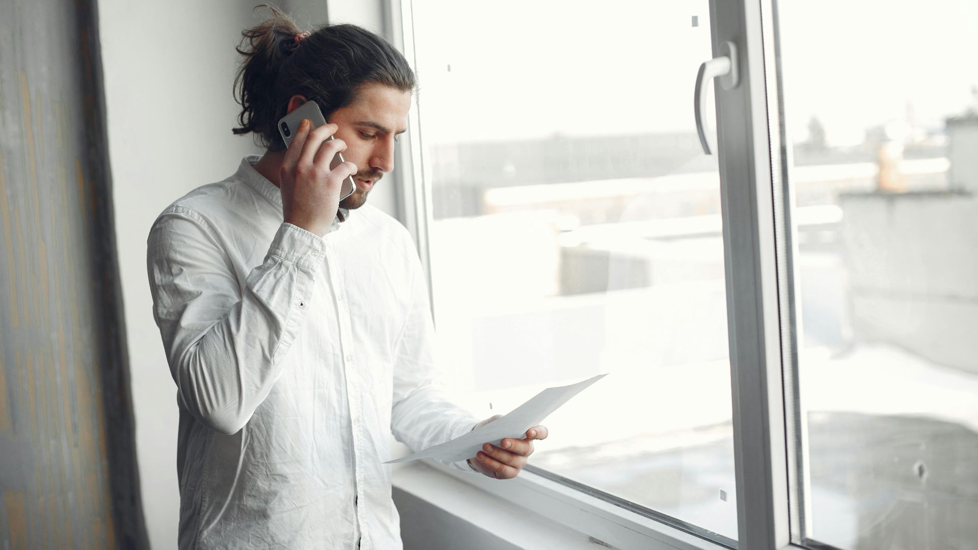 Young man in white shirt, on phone call holding a document, standing by a large window.