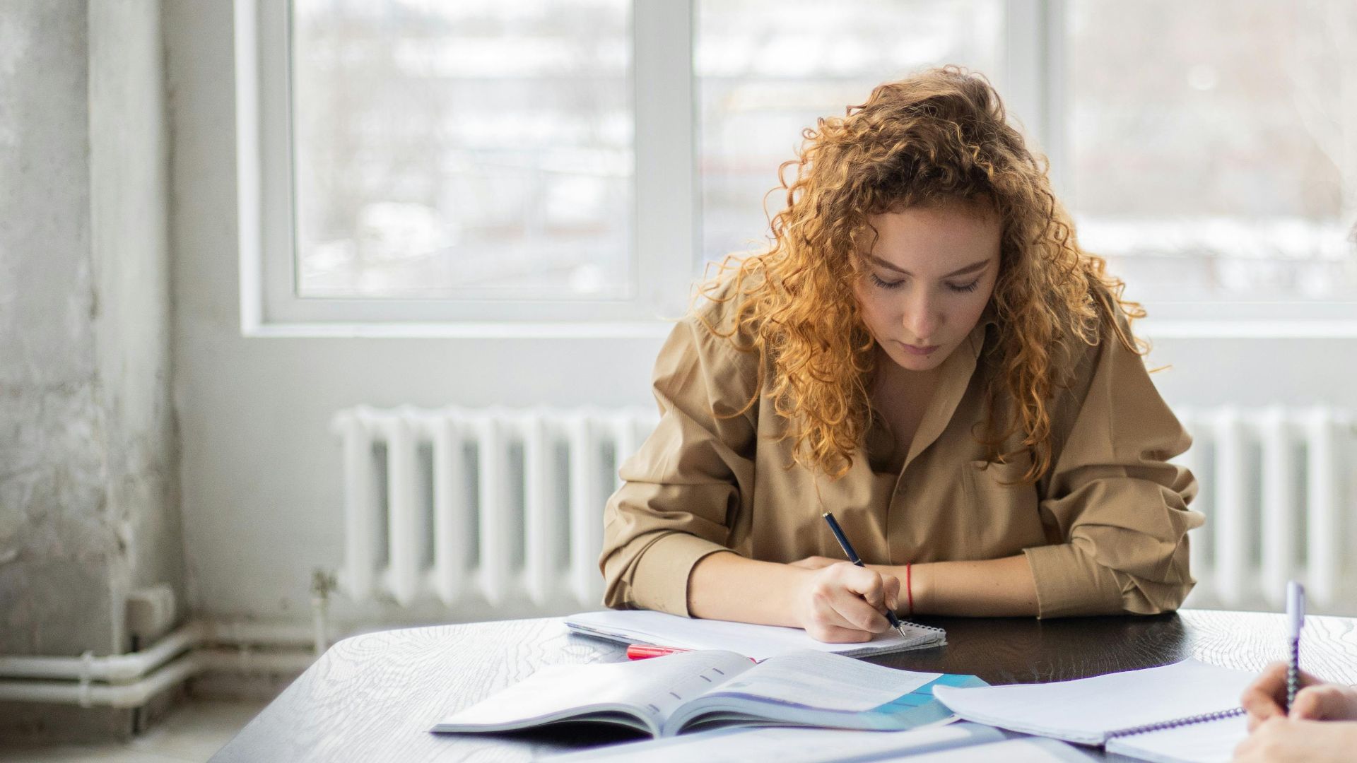 Focused students in casual clothes sitting at table with books and taking notes in notebook in light workspace in daytime