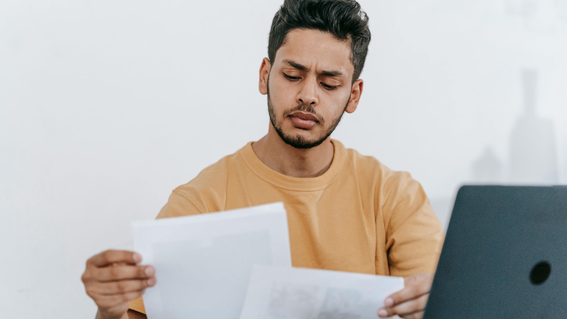 Attentive young bearded Hispanic male remote worker examining papers with results of project while working with laptop in home office