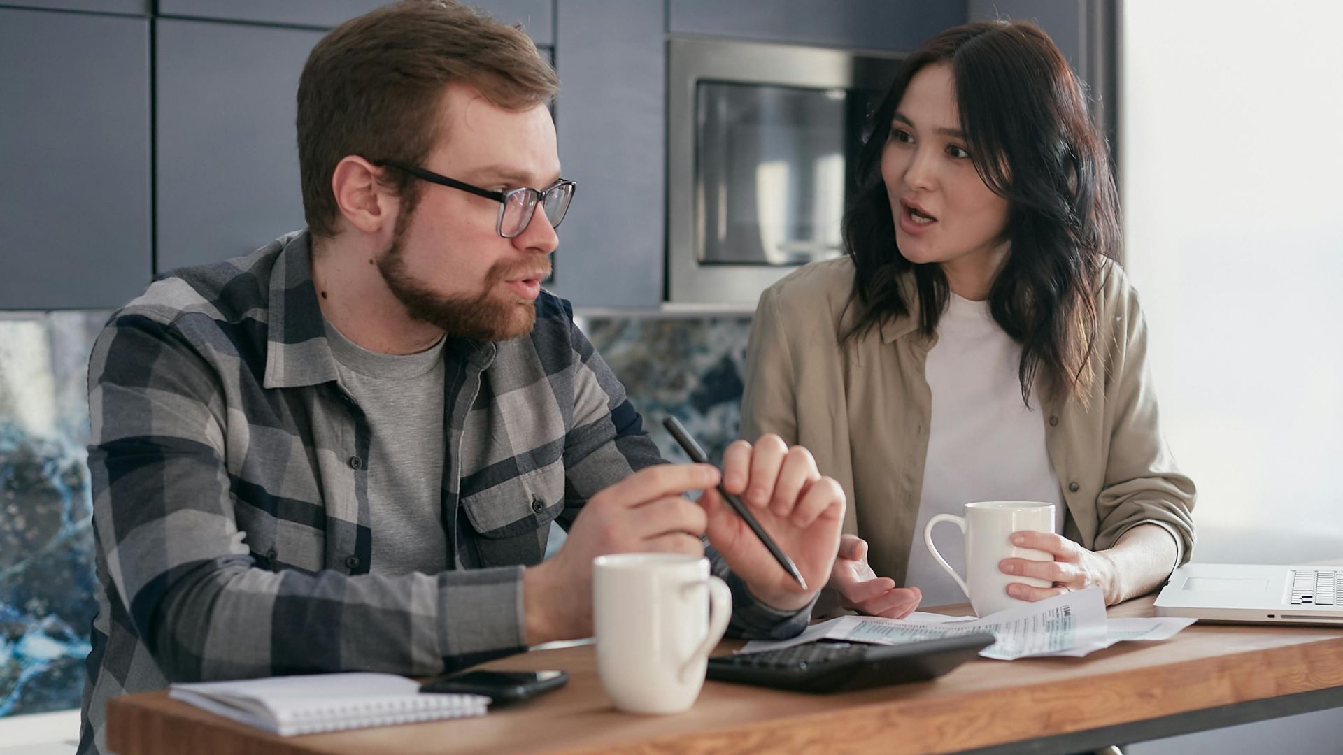 A couple managing finances with gadgets, documents, and coffee mugs at home.