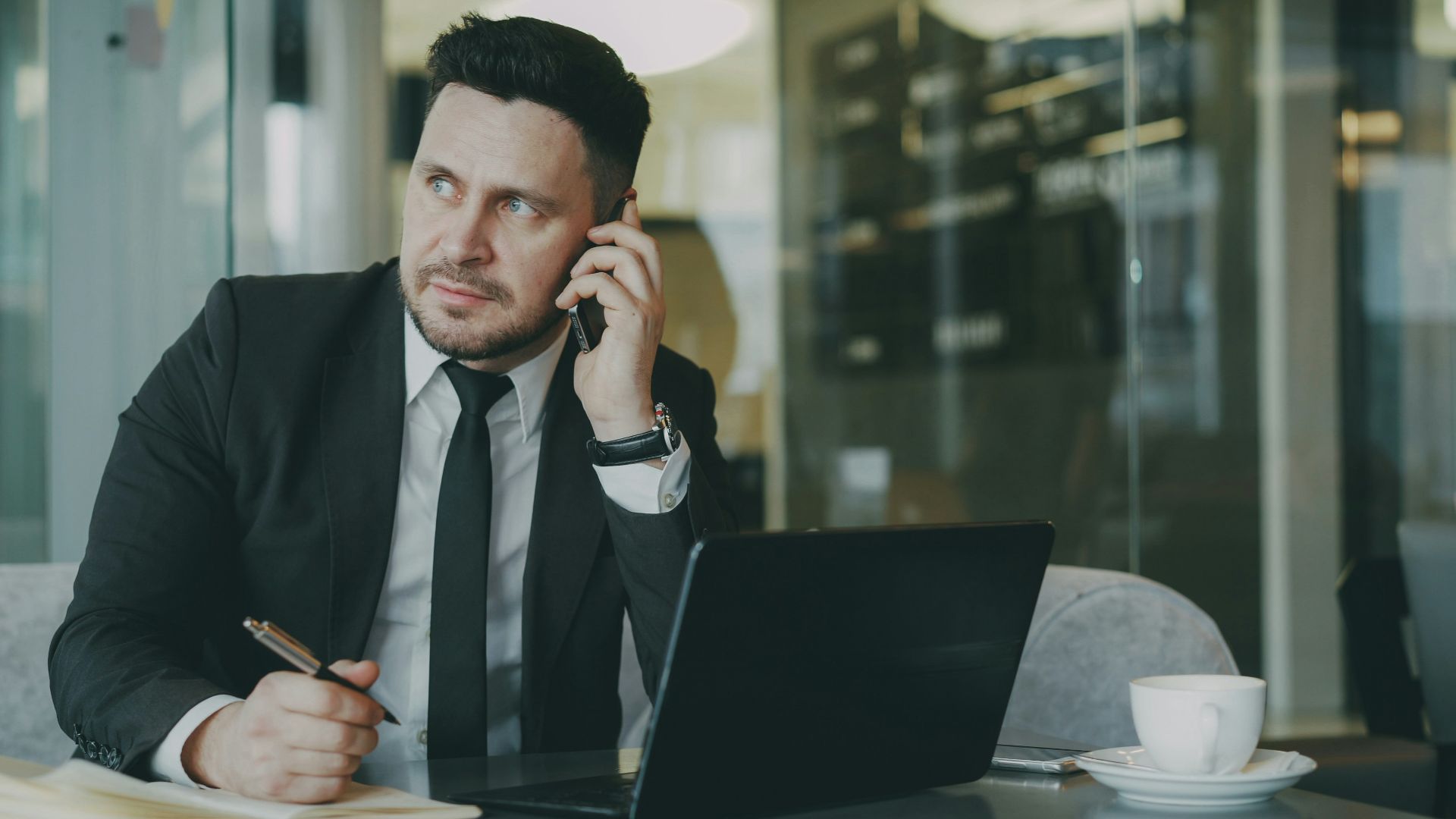Man in suit talking on phone at desk with laptop.