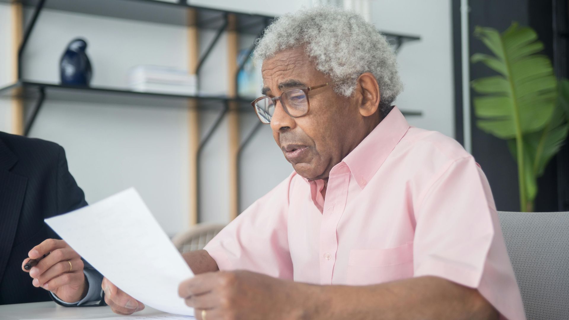Elderly man with glasses reading a document in a home office setting, reflecting focus and concentration.