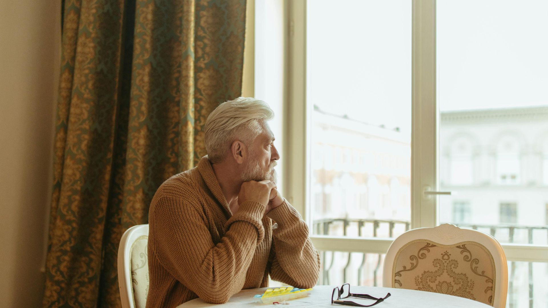 Elderly man in a cozy room gazing thoughtfully out the window during the day.