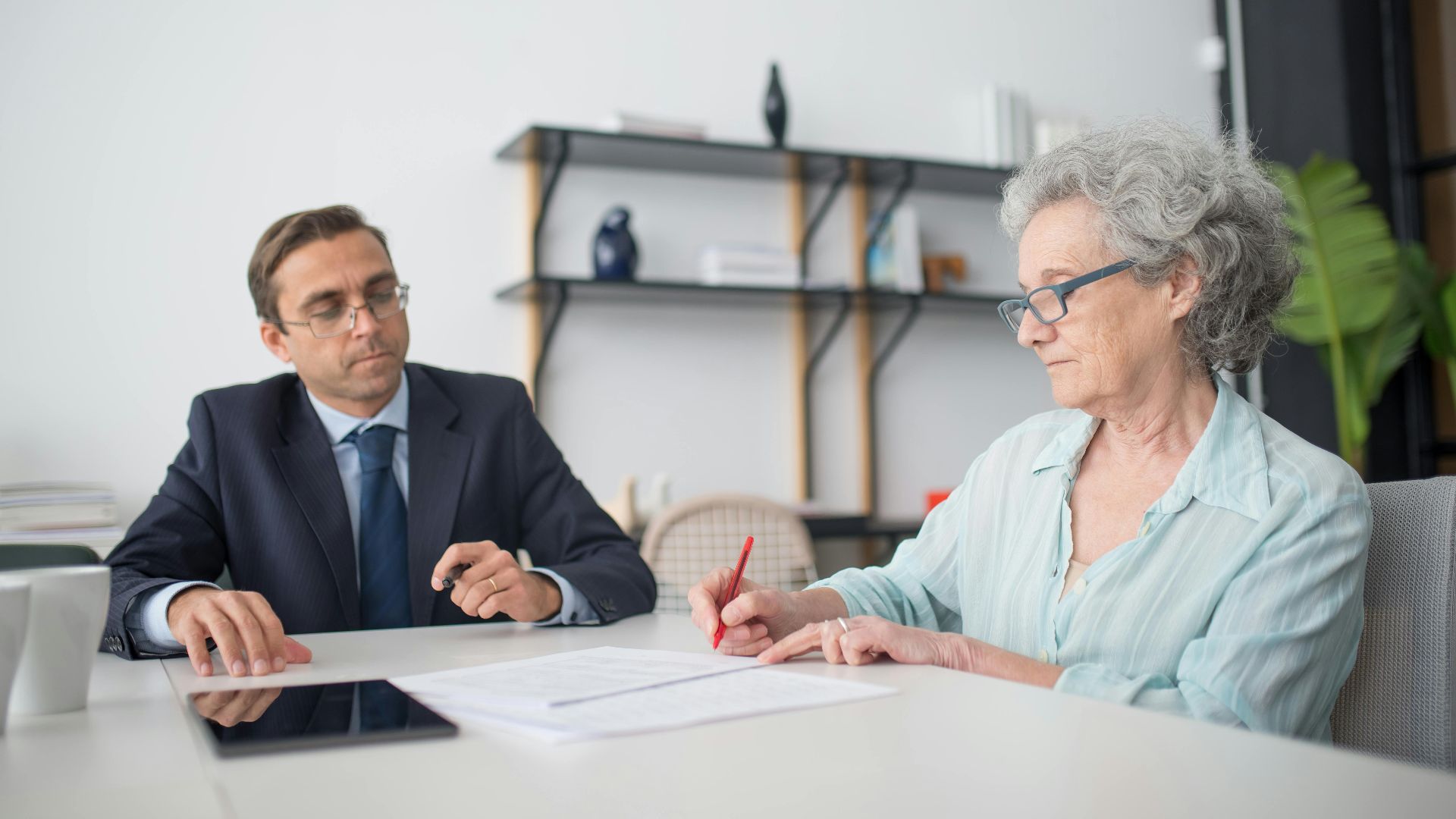 Elderly woman signing paperwork in modern office with consultant.