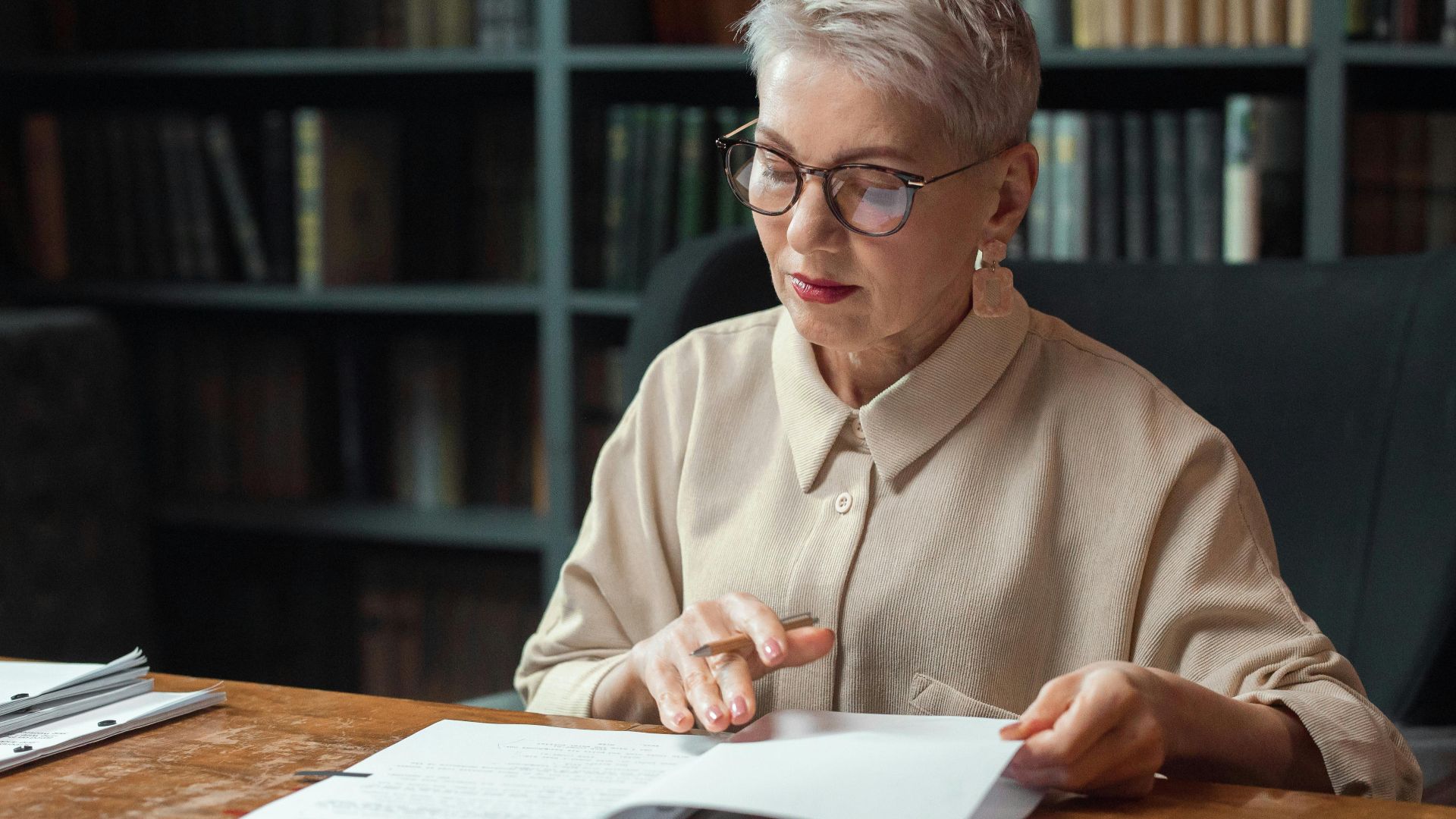 Elderly woman with glasses reading documents at a desk in a library.