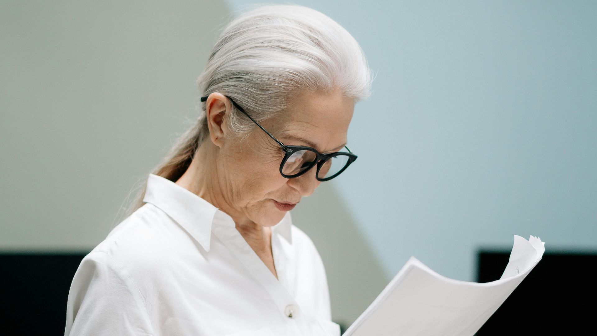 Elderly woman with eyeglasses reviews papers in modern office, surrounded by plants.
