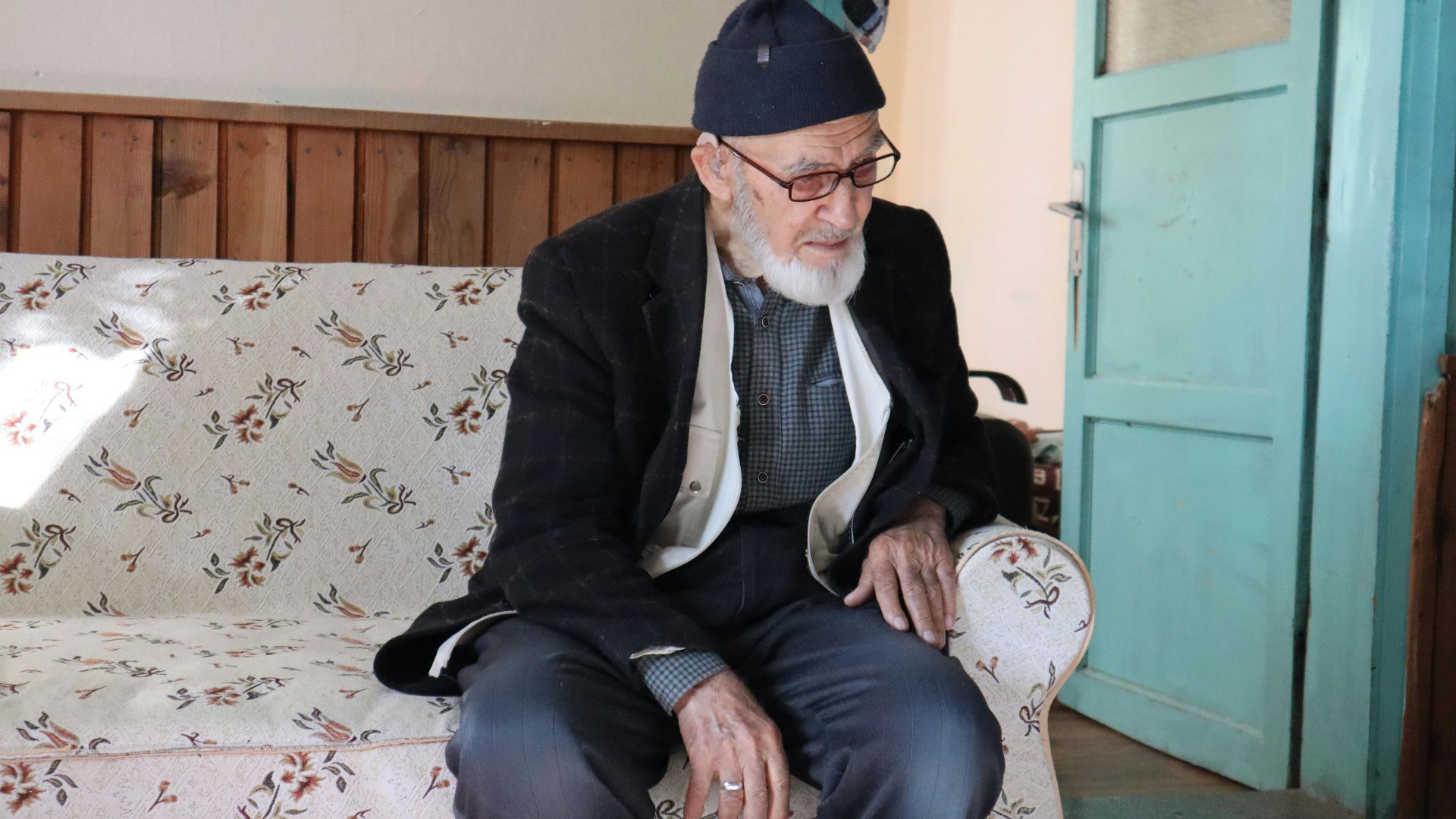 Elderly man sitting on a floral sofa indoors, enjoying a moment of relaxation and contemplation.