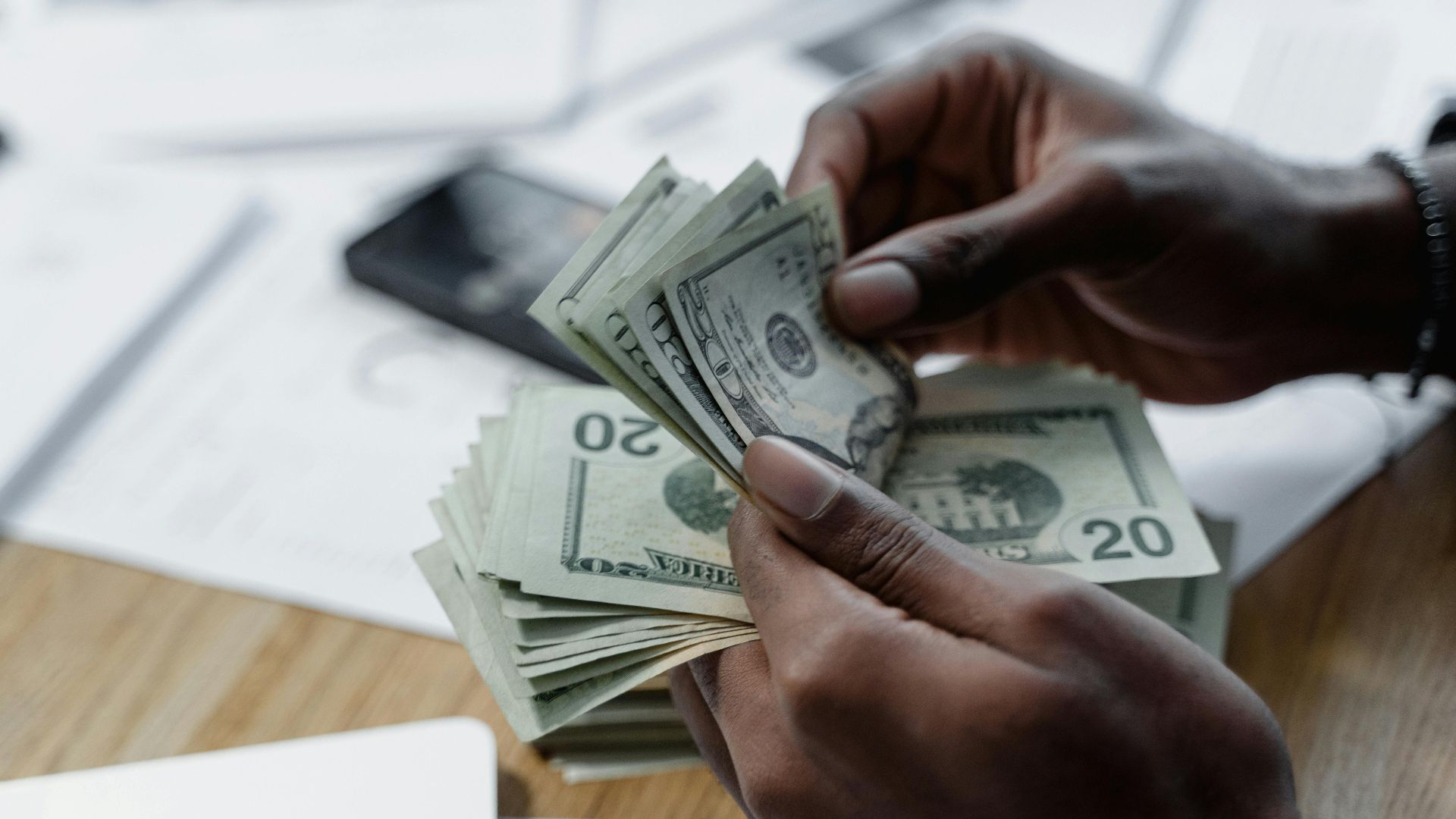A close-up of hands counting US dollar bills on a wooden table with documents and a smartphone nearby.