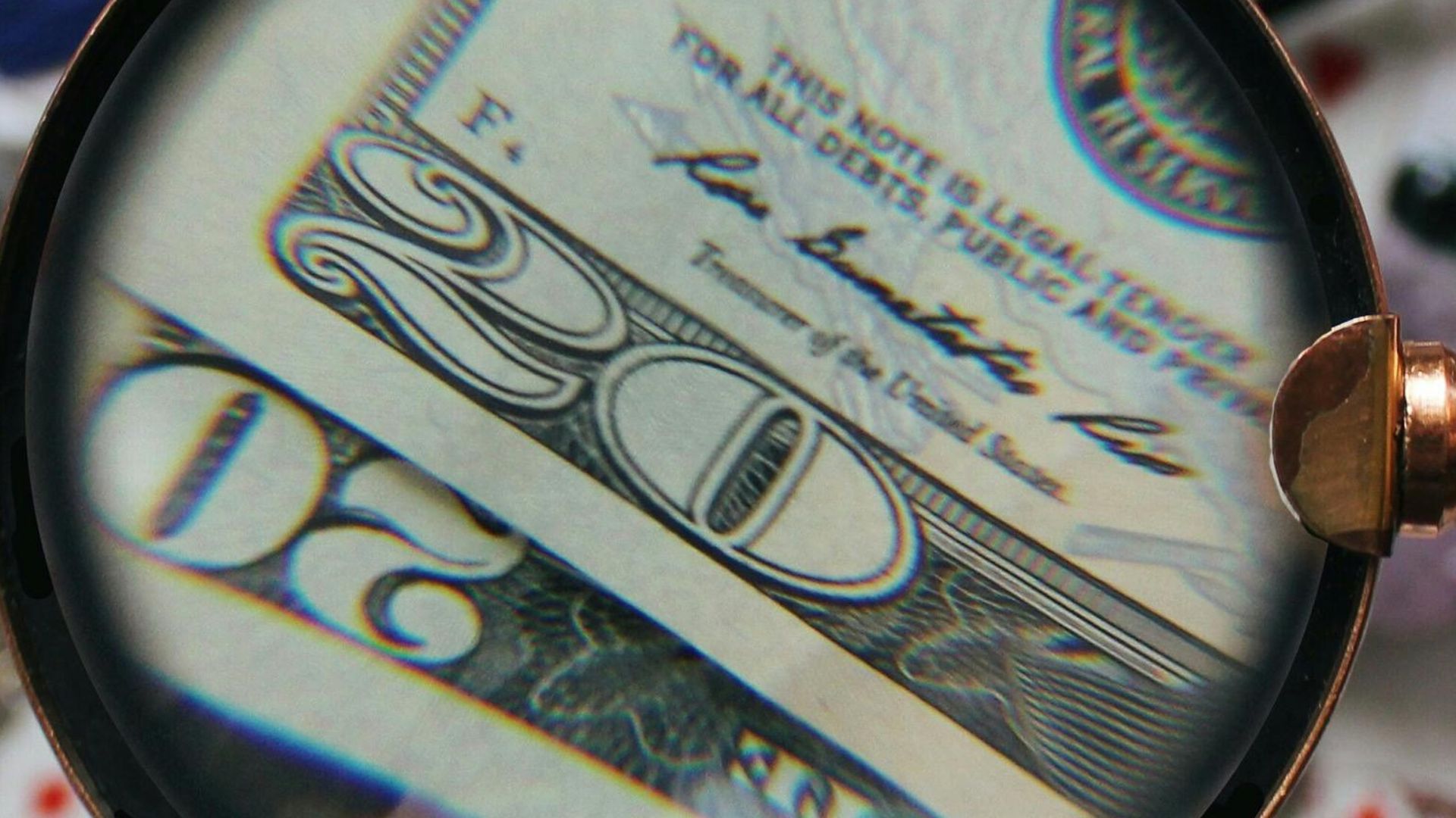 A hand holding a magnifying glass focusing on a twenty dollar bill amidst various scattered objects.