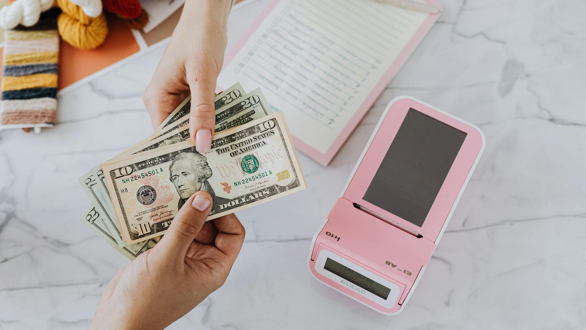 Close-up of hands exchanging US dollars with a pink calculator on a marble surface.