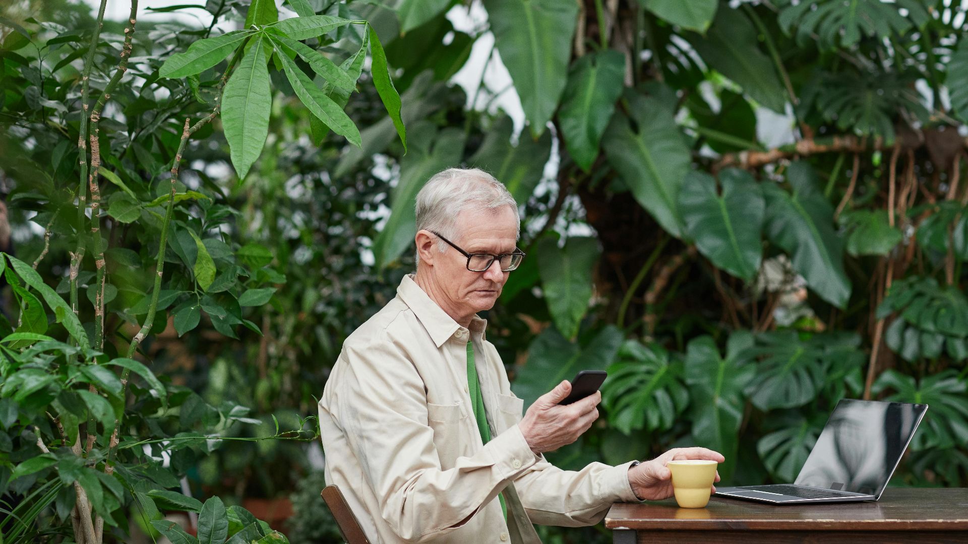 Elderly man engaged in remote work with laptop and smartphone surrounded by lush greenery.