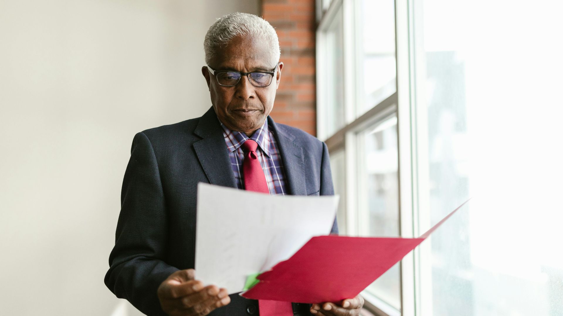 Elderly man in business attire examining papers by a window in an office setting.