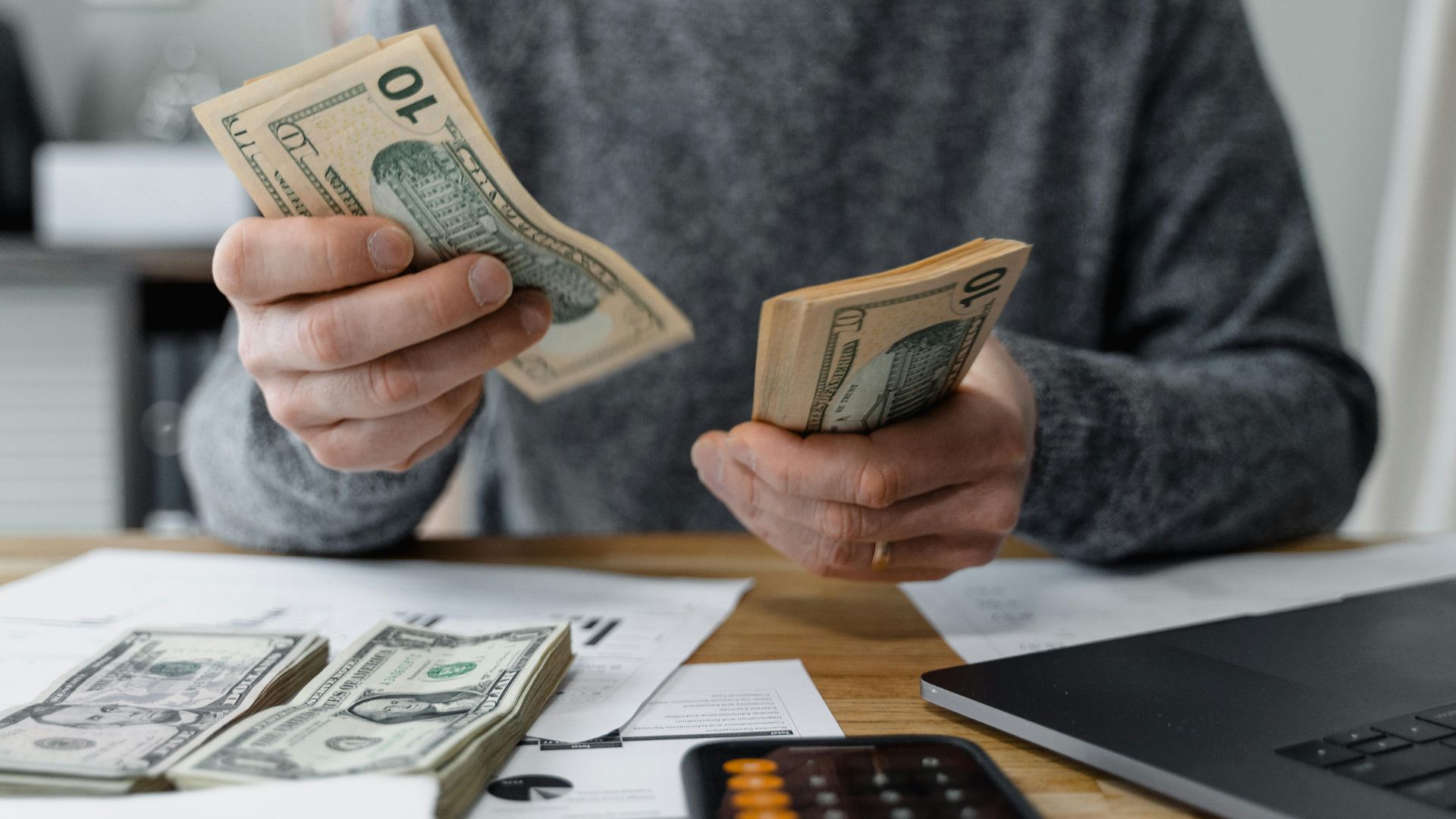 Close-up of person counting cash with a calculator and paperwork on a desk.