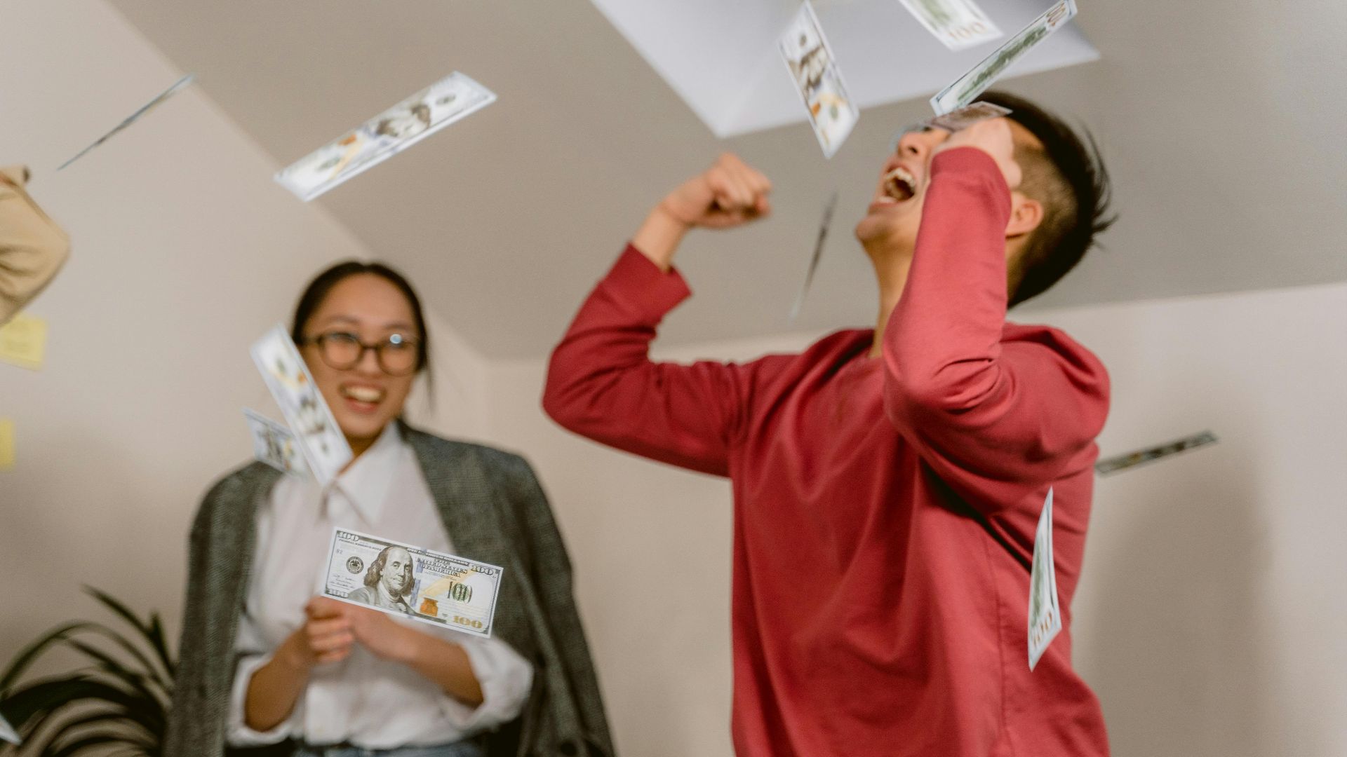 Two young adults in a room celebrating with falling dollar bills, expressing excitement and joy.