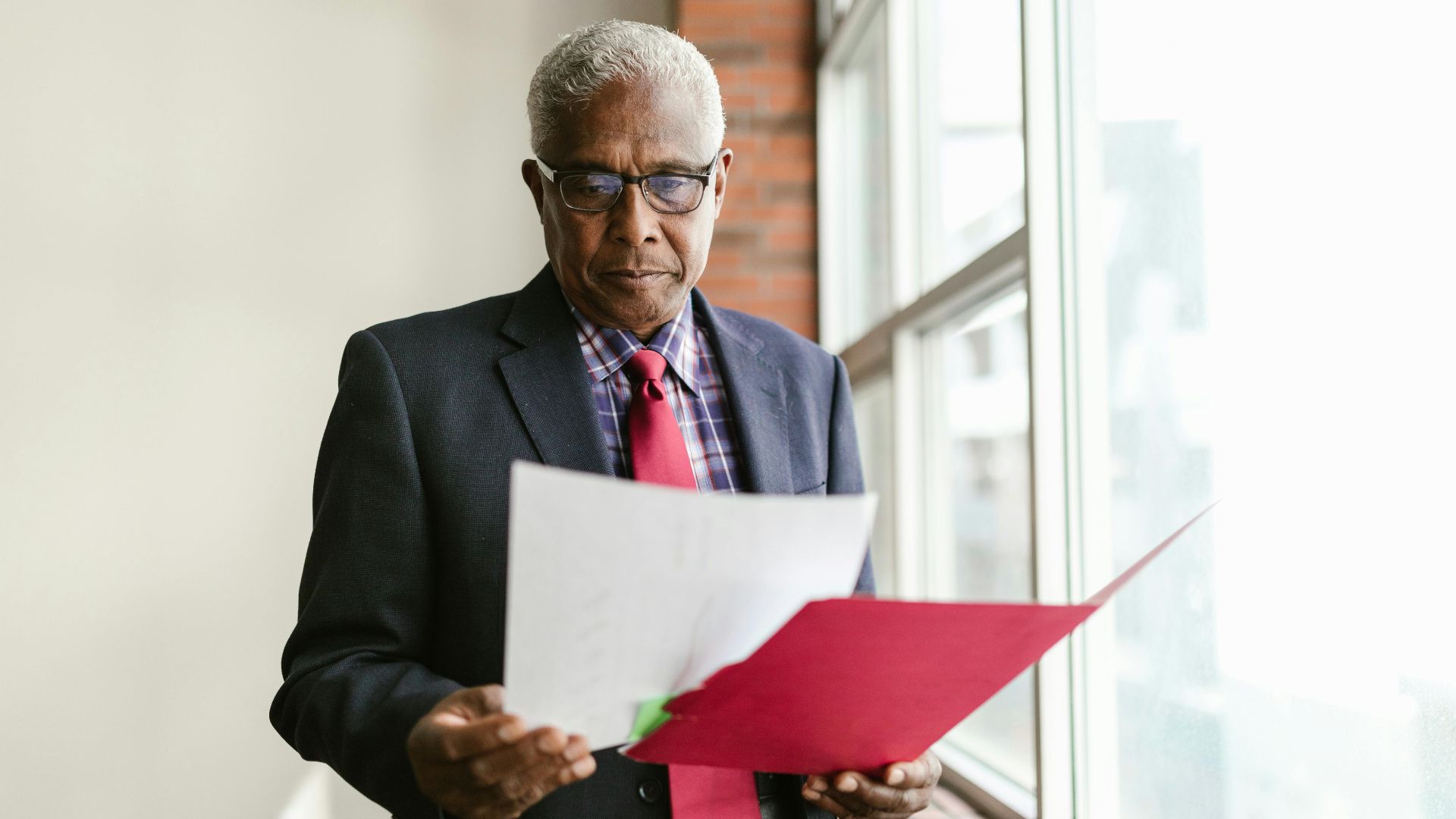 Elderly man in business attire examining papers by a window in an office setting.