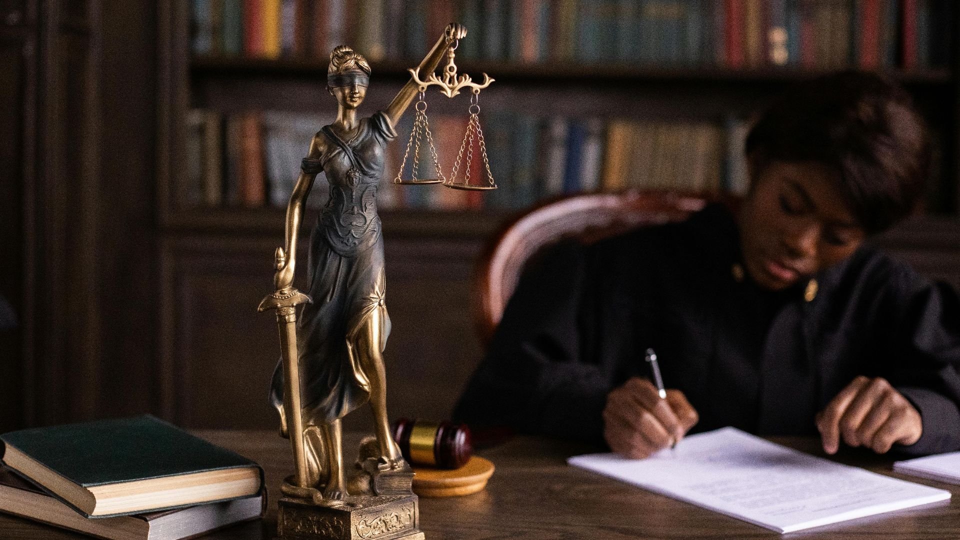 A focused judge writing on documents beside a Lady Justice statue in an office.