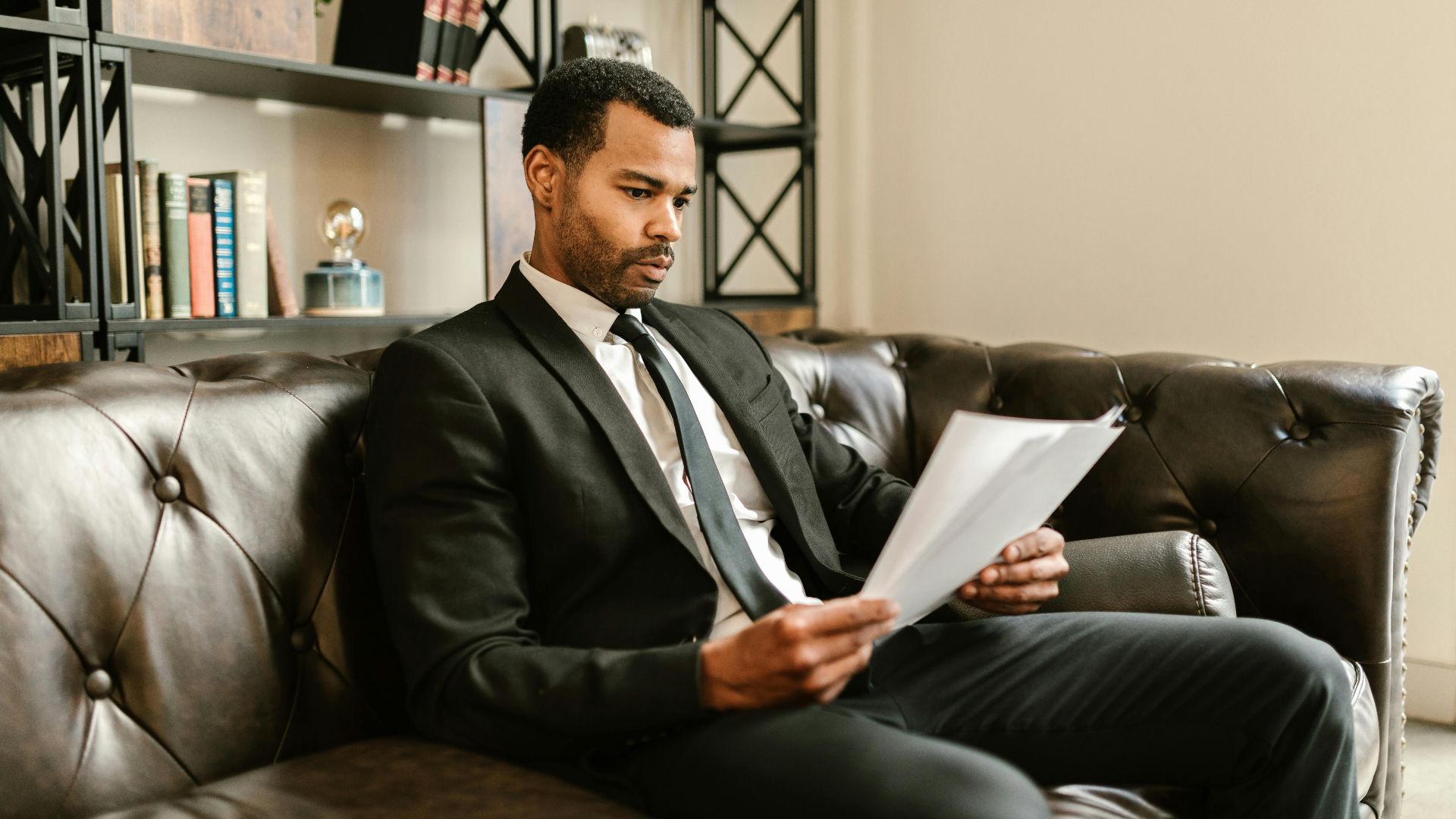 Businessman in suit reading papers on a leather sofa in a modern office setting.