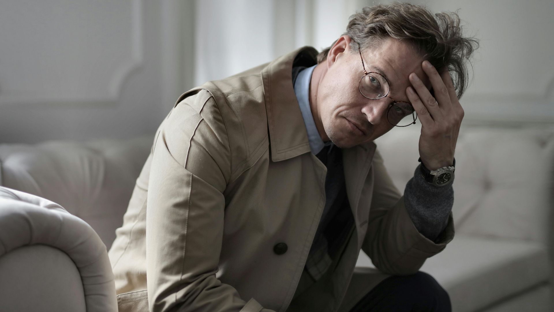 Pensive adult brown haired man in eyeglasses and trench sitting on couch and touching head while looking at camera