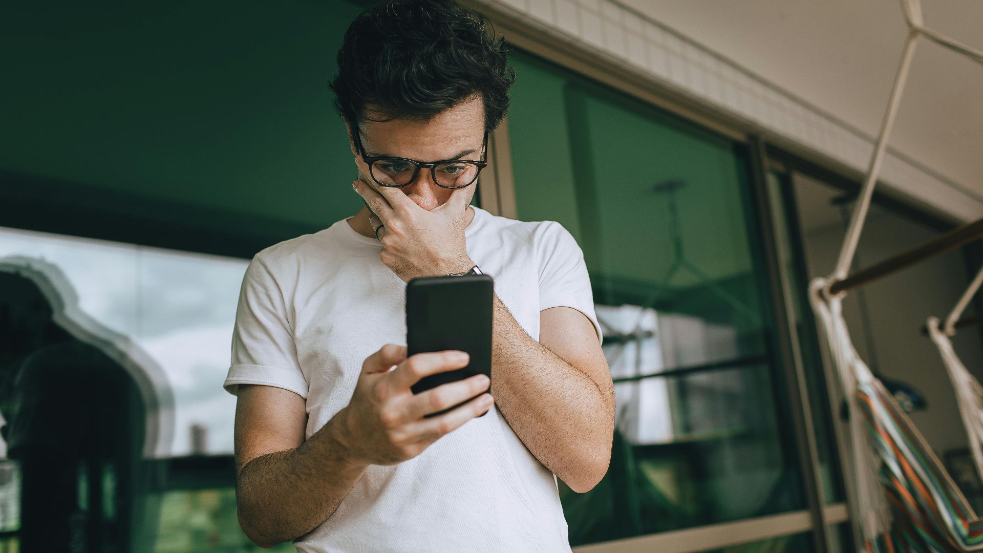 Man with eyeglasses focused on smartphone indoors, captured in a contemplative moment.
