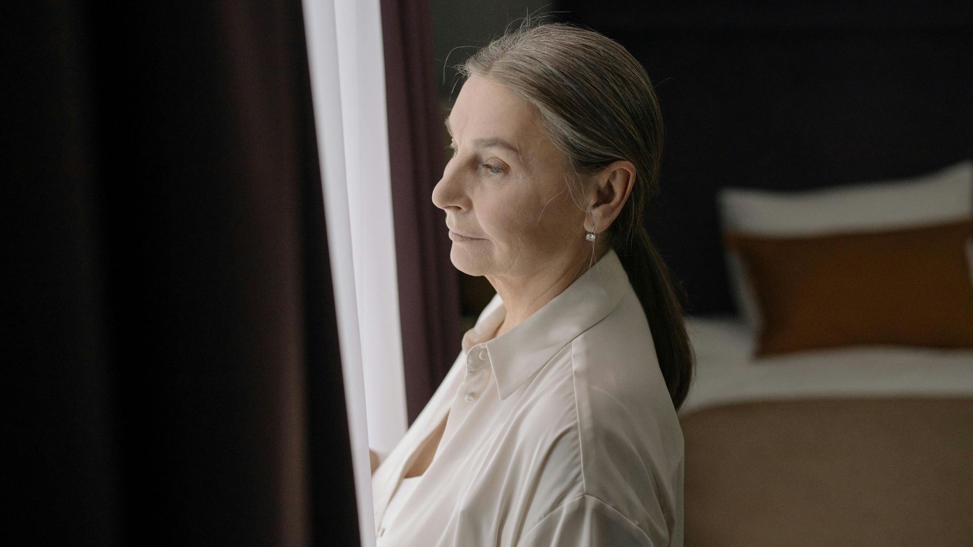 Senior woman with grey hair in profile, gazing out a window, conveying peaceful reflection.