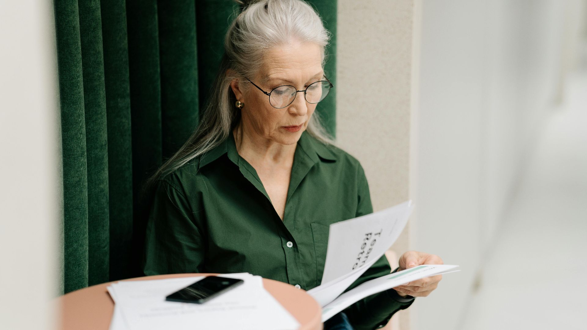 Elderly woman with gray hair reading papers at a small table indoors, focused and thoughtful.