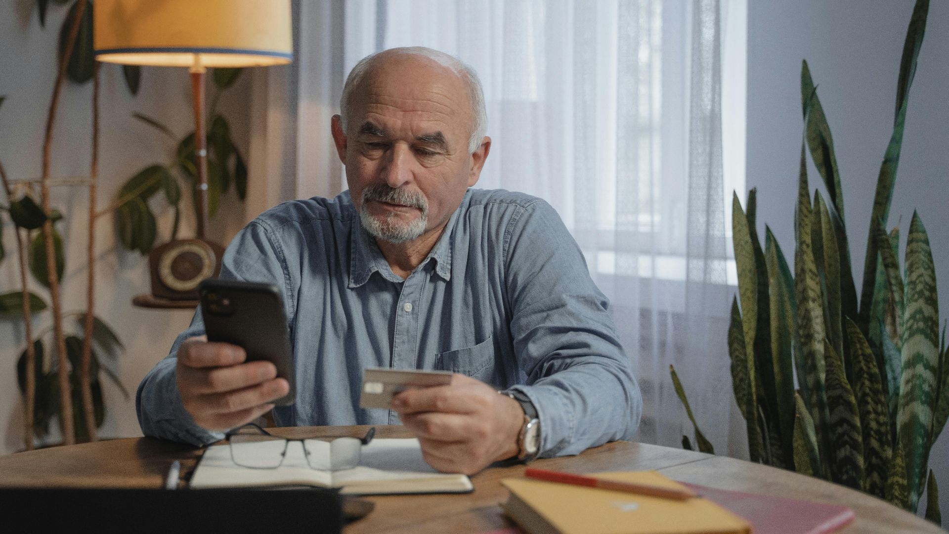 Elderly man using smartphone for online shopping at home, holding credit card near desk plants.