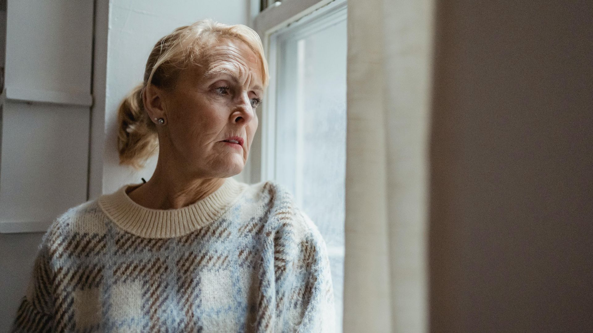 Contemplative senior female wearing warm sweater sitting on windowsill and looking away in sad thoughts